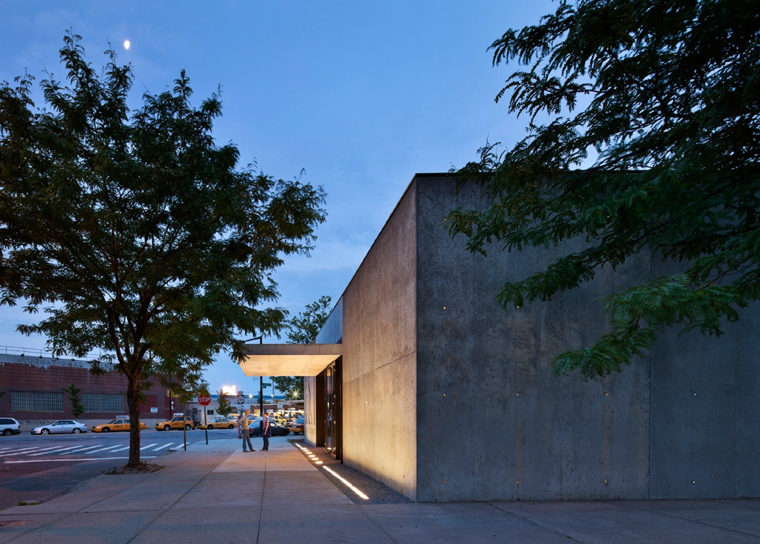 MoMA PS1 Entrance Building by Andrew Berman Architect — The entrance pavilion presents itself as a monolithic concrete volume at dusk, its board-formed walls punctuated by embedded glass rods that glow as...