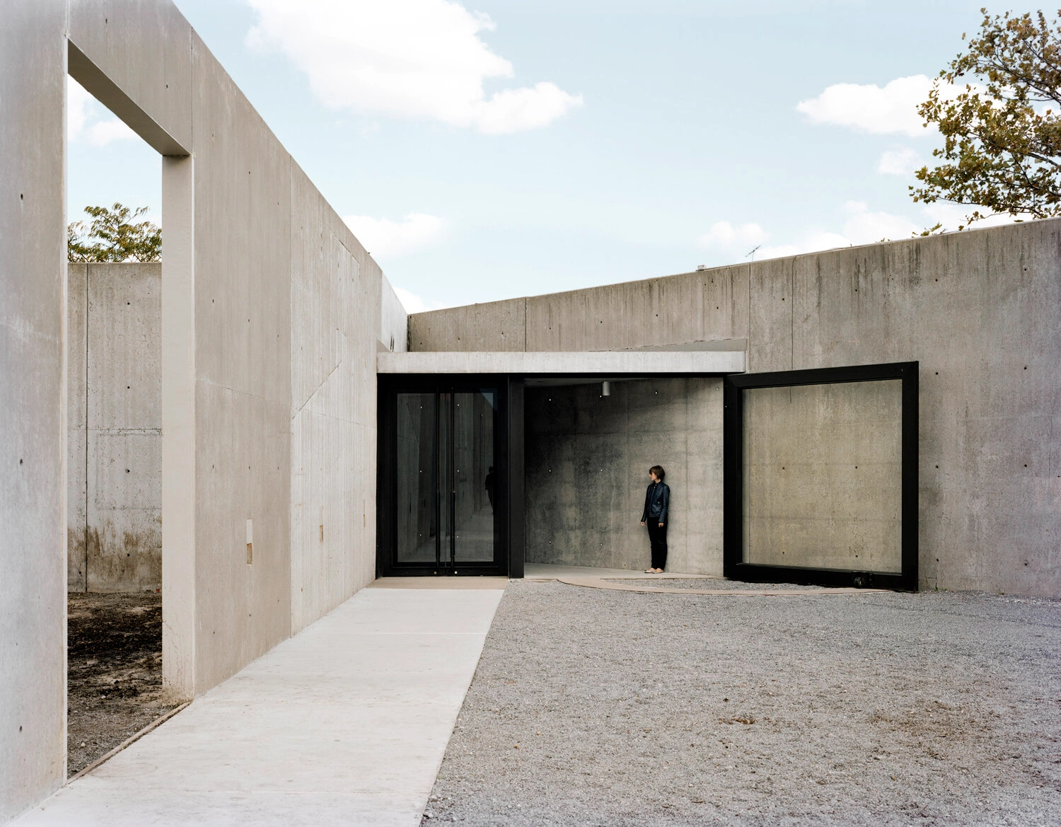 MoMA PS1 Entrance Building by Andrew Berman Architect — The entrance courtyard frames a monolithic concrete pavilion with board-formed walls displaying visible formwork texture and weathering marks.