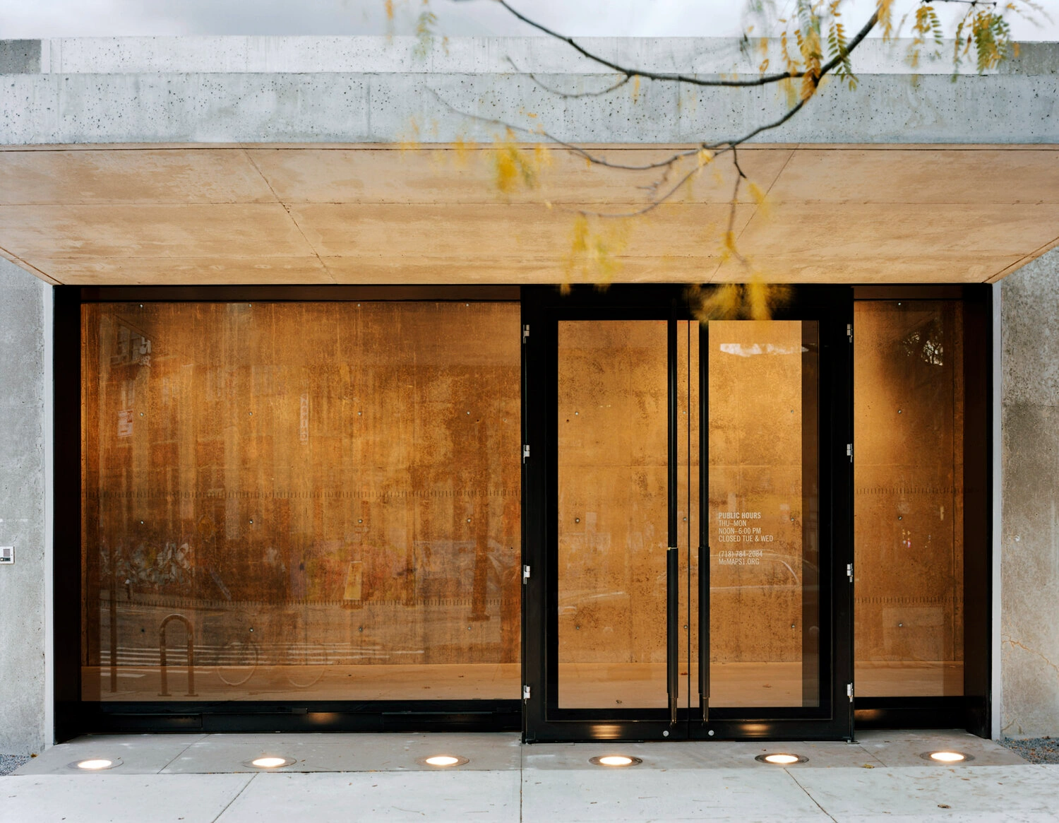 MoMA PS1 Entrance Building by Andrew Berman Architect — The entrance threshold presents a monolithic concrete facade punctuated by three black-framed glass doors with rust-stained glass panels.