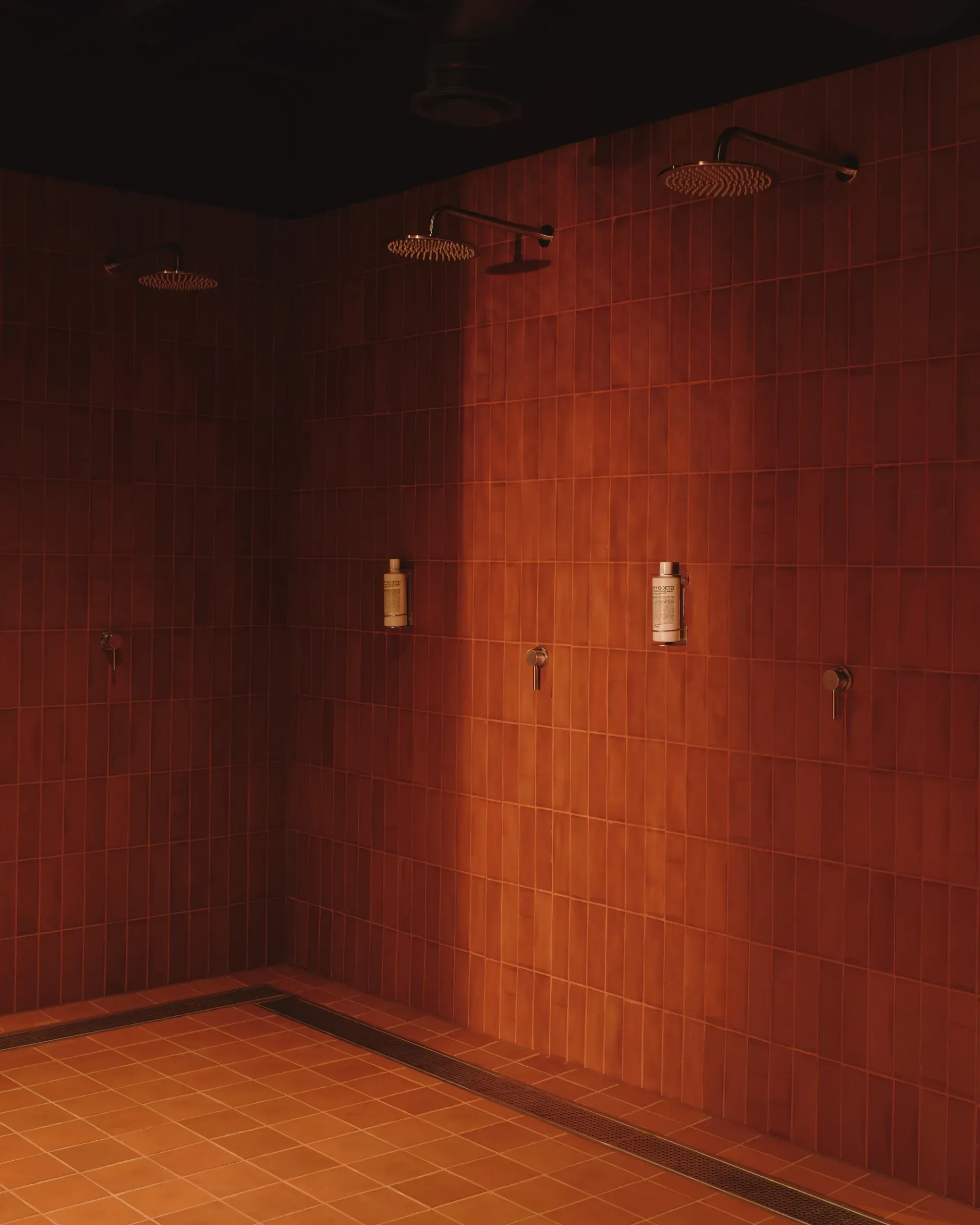 Arc Canary Wharf by Cake Architecture — The shower area features walls clad entirely in vertically-aligned terracotta tiles that shift in tone from deep rust to warm orange under directed lighting.