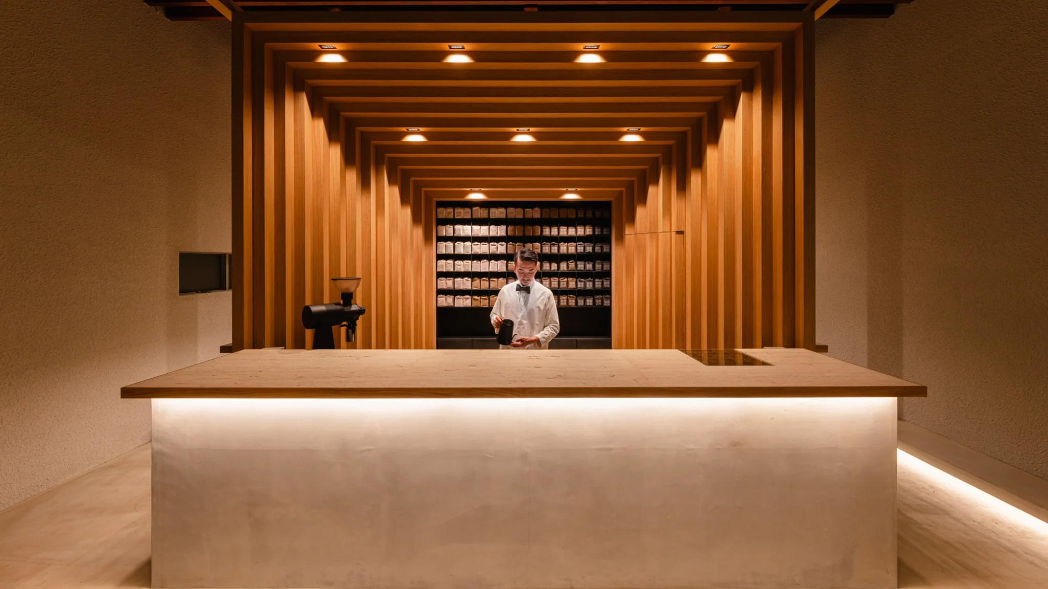 Koffee Mameya Kakeru by Fourteen Stones Design — A Tokyo coffee counter captured from the street threshold, framing the barista centrally behind a minimalist white concrete service counter.