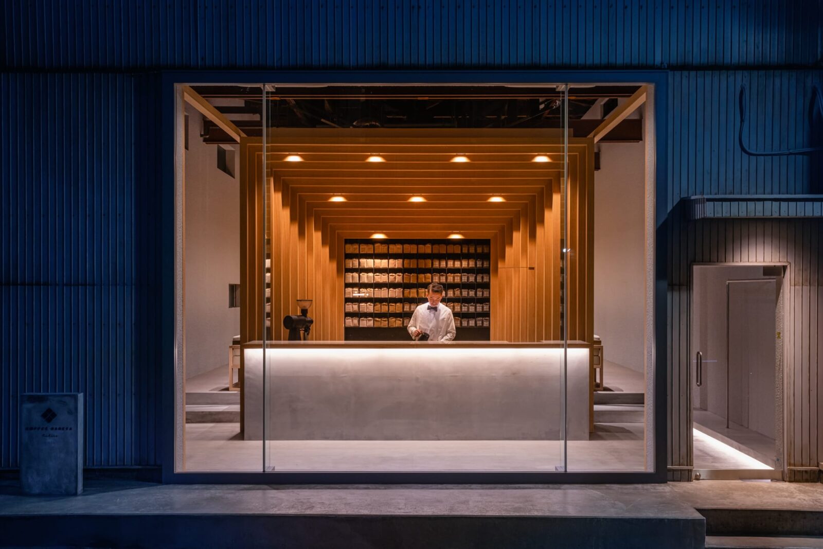 Koffee Mameya Kakeru by Fourteen Stones Design — A corrugated metal warehouse facade in charcoal blue frames a full-height frameless glass storefront.