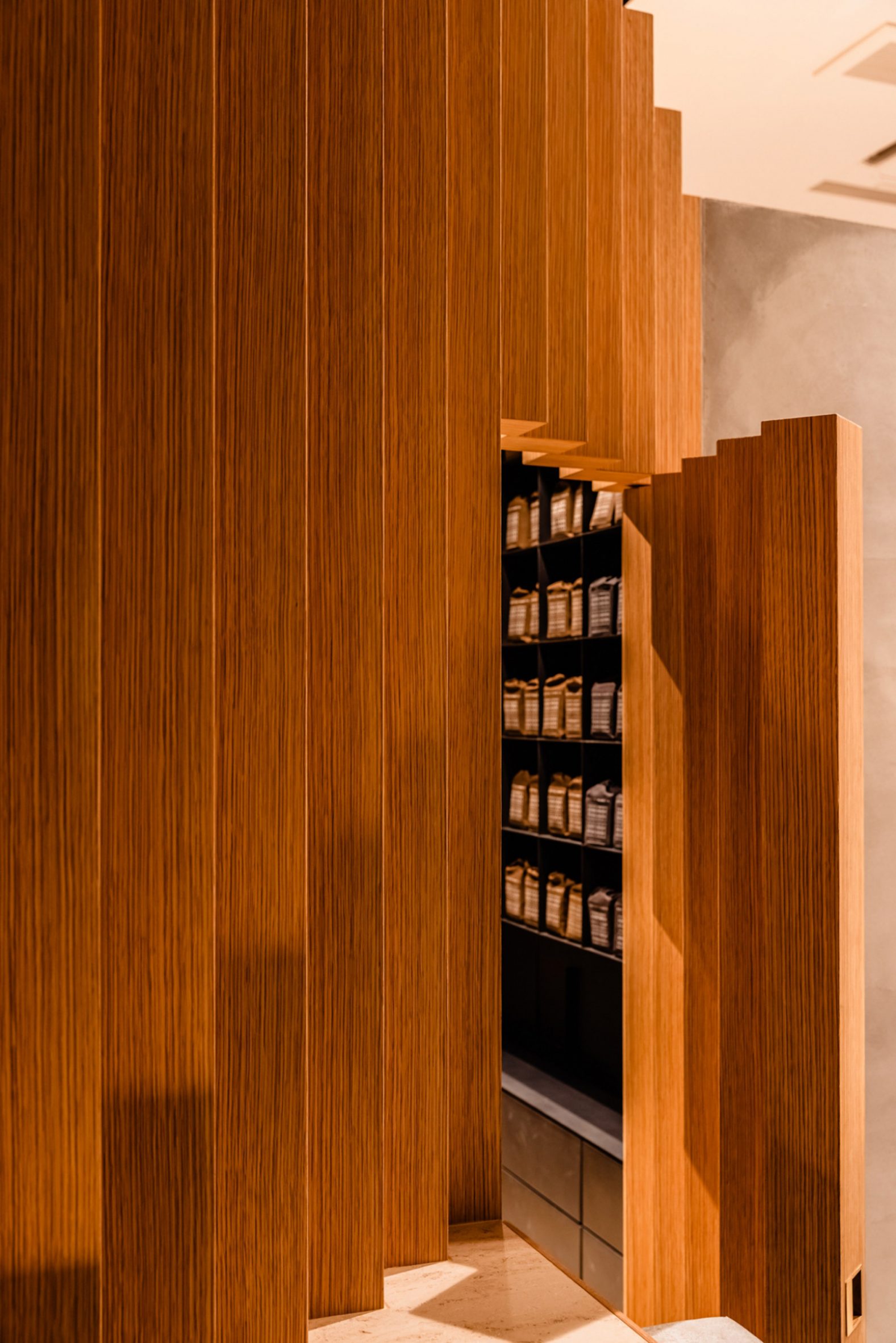 Koffee Mameya Kakeru by Fourteen Stones Design — A concealed shelving system materializes behind vertical timber slats, revealed through a narrow passage between two fluted wood panels.