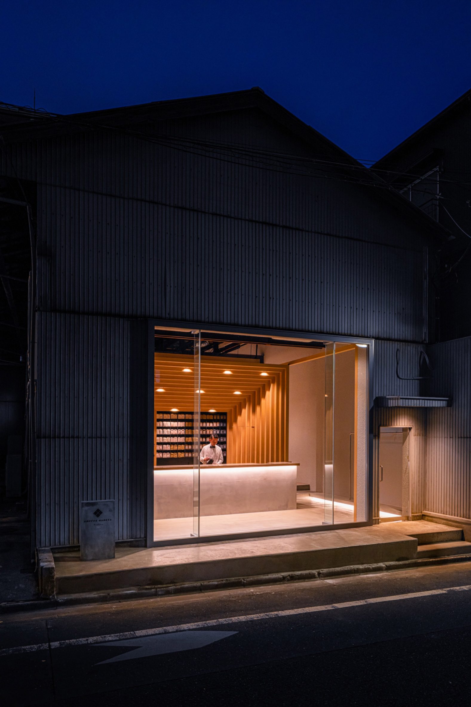 Koffee Mameya Kakeru by Fourteen Stones Design — A converted warehouse storefront photographed at dusk features a dark corrugated metal exterior shell contrasting sharply with a full-height frameless glass...