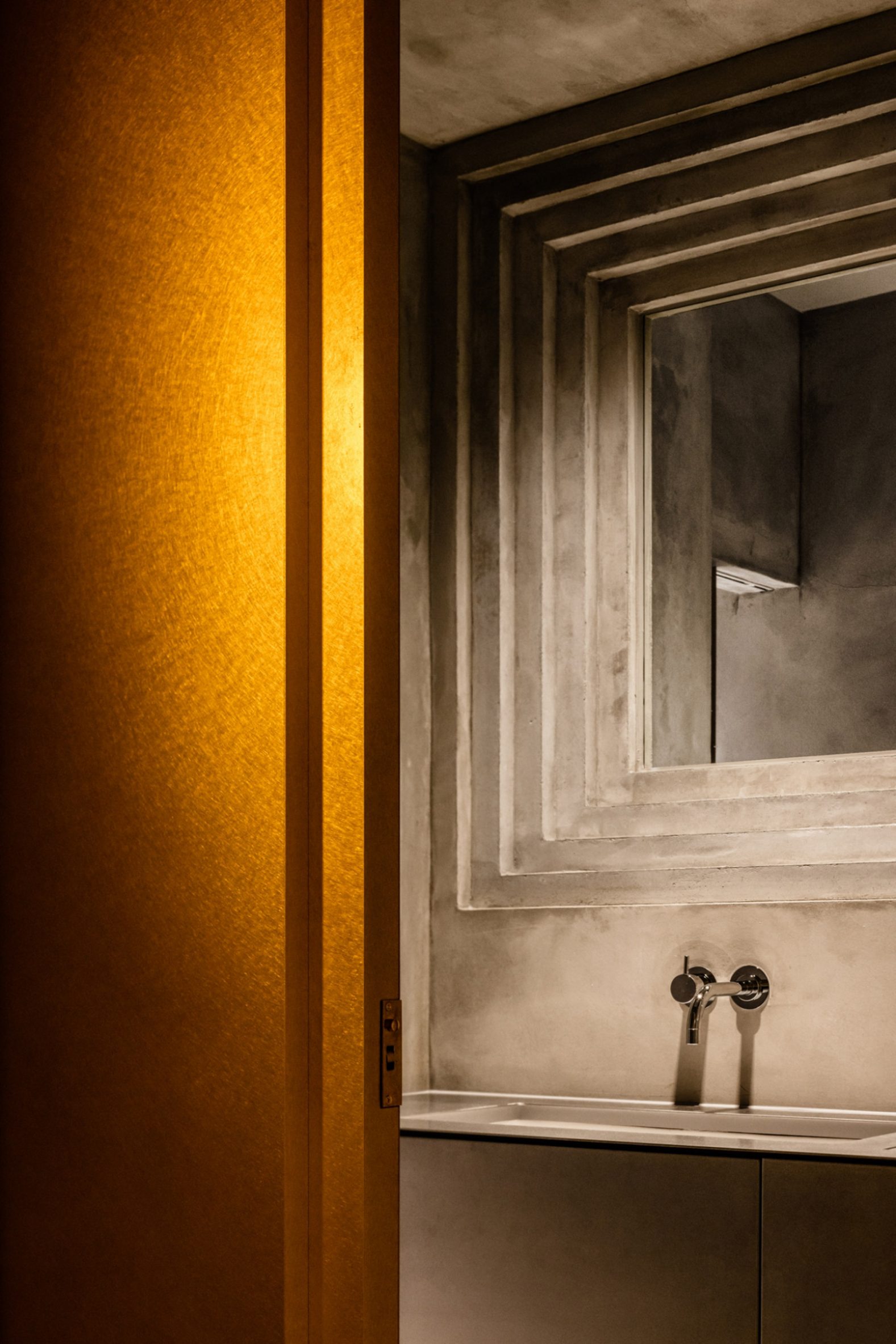 Koffee Mameya Kakeru by Fourteen Stones Design — A bathroom detail captured from the threshold, the golden-orange door frame dominates the left plane while a recessed mirror with nested rectangular frames...