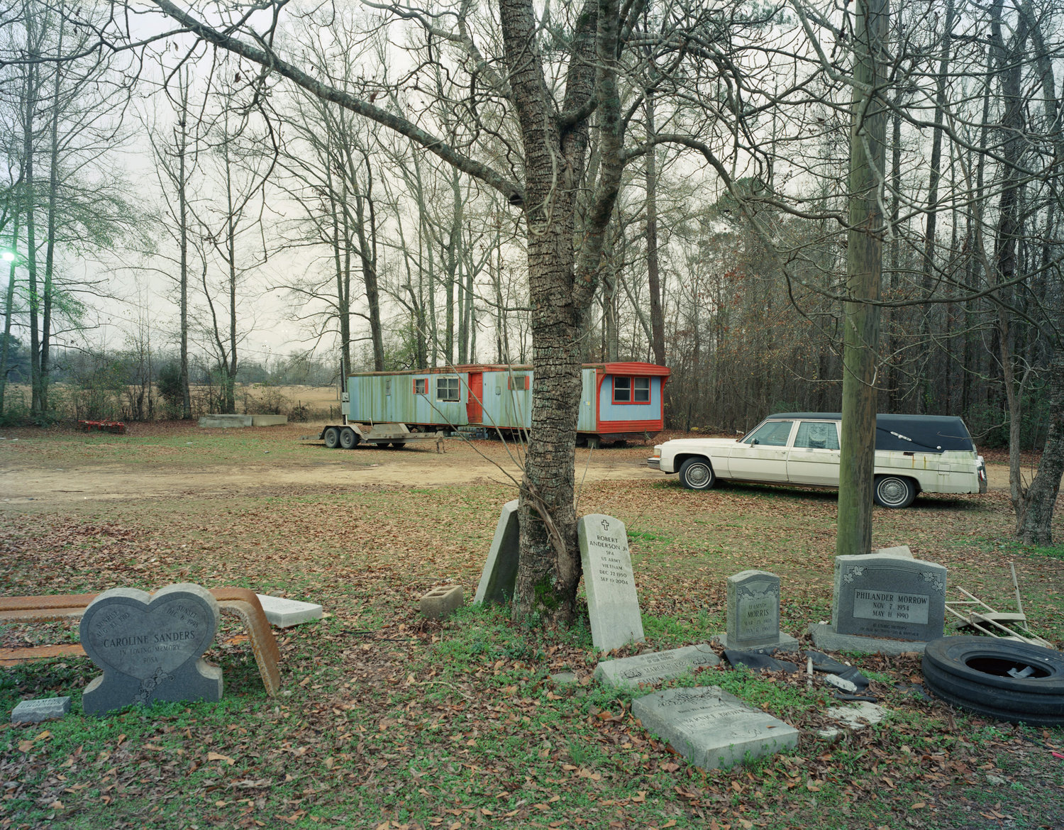 Funeral Homes of the American South by Timothy Hursley — A clearing in bare woodland contains two mobile trailers and parked automobiles positioned among grave markers and a discarded tire.