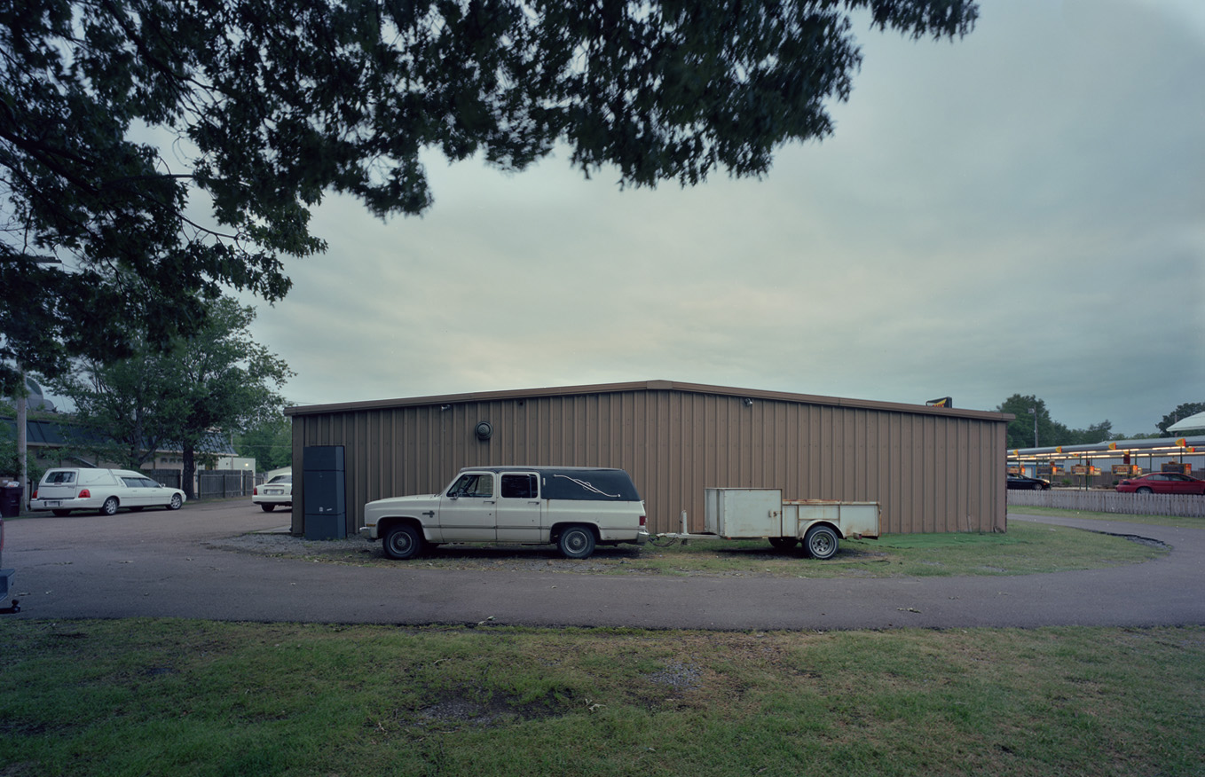 Funeral Homes of the American South by Timothy Hursley — A cream-colored SUV with matching utility trailer sits parked against a corrugated metal industrial building under overcast sky.