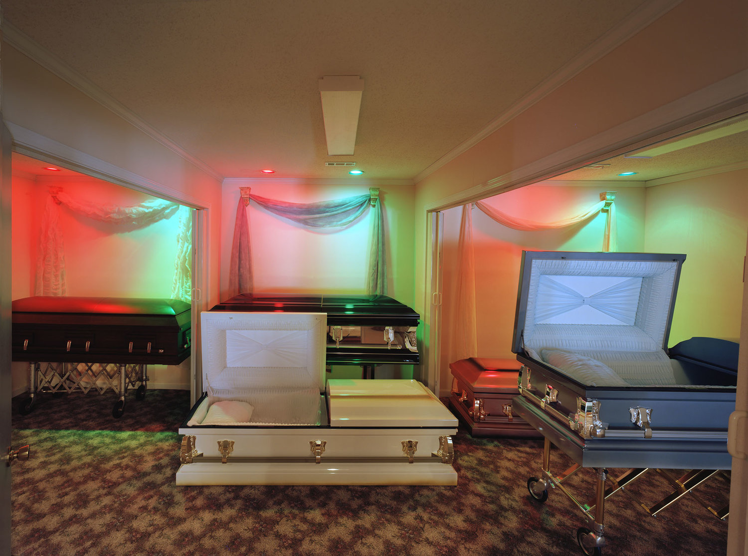 Funeral Homes of the American South by Timothy Hursley — A showroom display of caskets arranged in a climate-controlled interior with chromatic theatrical lighting.