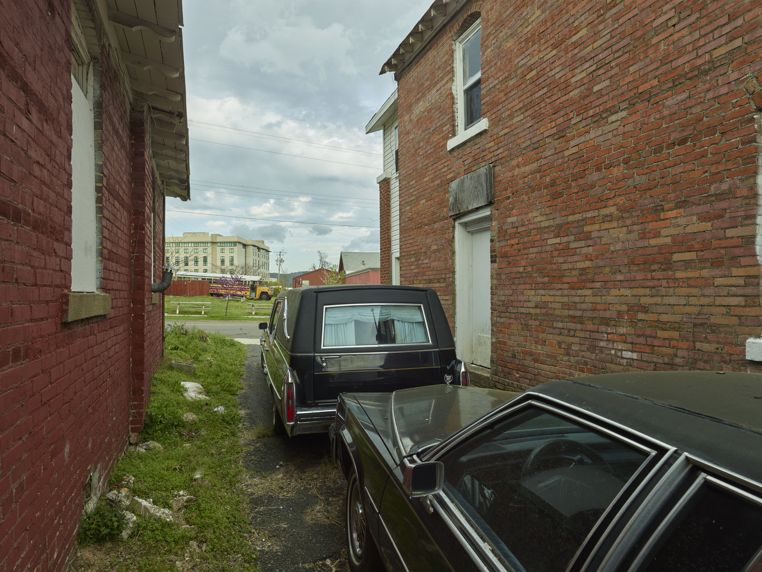 Funeral Homes of the American South by Timothy Hursley — A narrow passage between two brick buildings frames a black hearse parked in the alley, its chrome trim and rear window catching gray daylight.