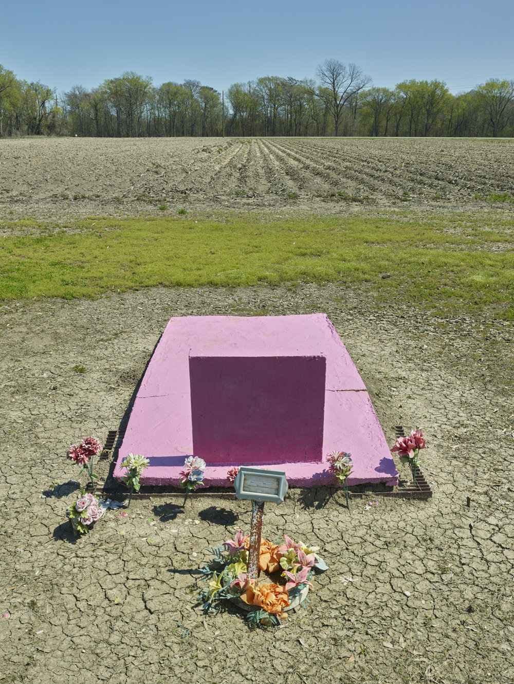 Funeral Homes of the American South by Timothy Hursley — A pink concrete vault sits centered in an expansive agricultural field, surrounded by cut flowers and a small digital display device.