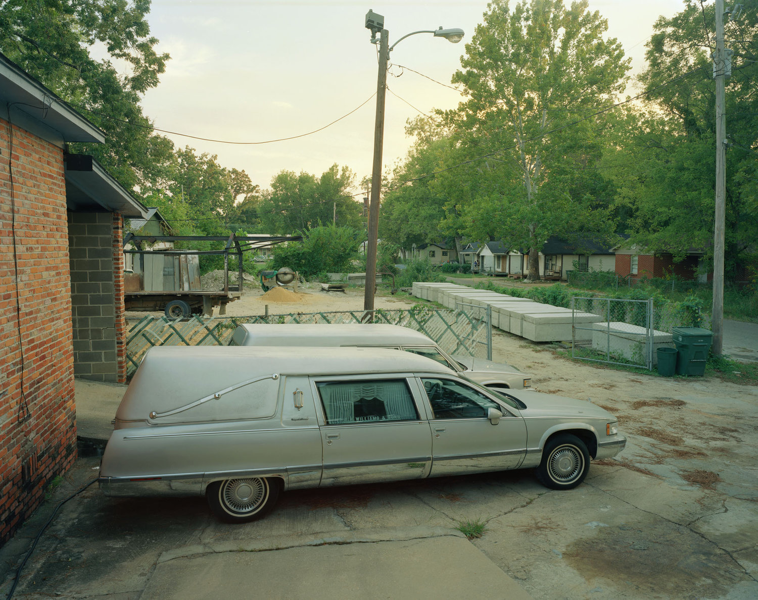 Funeral Homes of the American South by Timothy Hursley — A silver Cadillac hearse sits parked on a concrete pad beside a brick building, its elongated profile dominating the foreground.