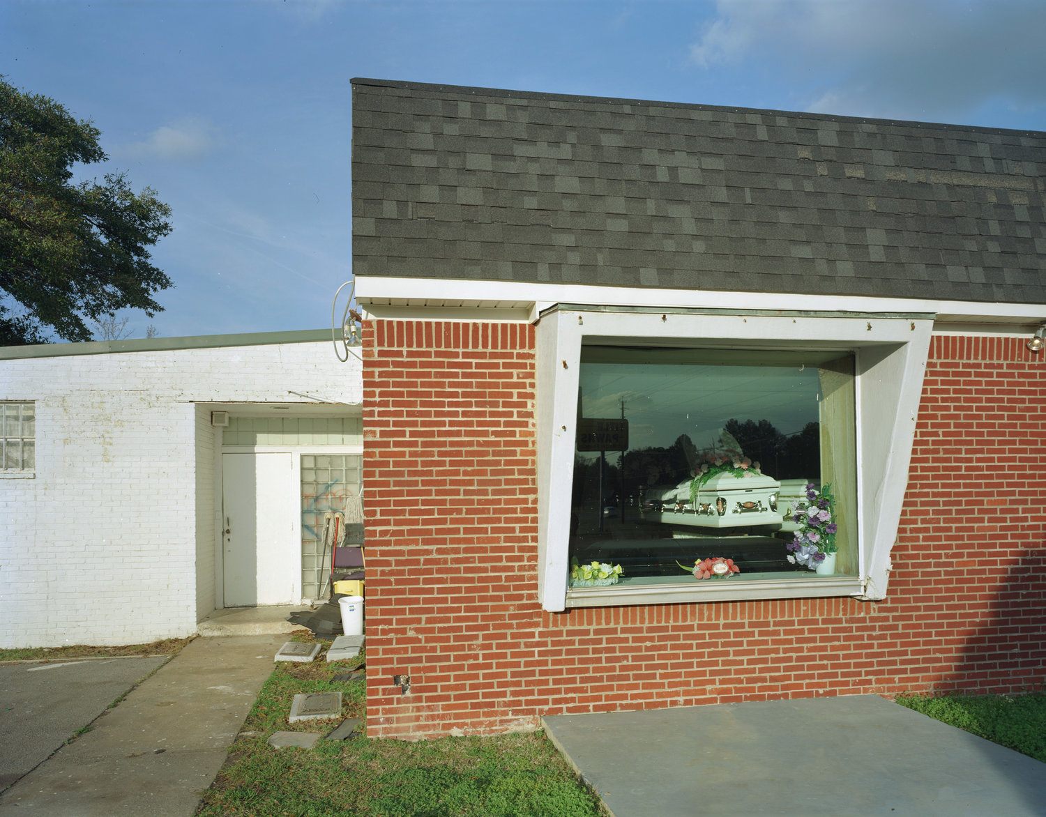 Funeral Homes of the American South by Timothy Hursley — A brick funeral home's display window frames a casket arrangement with floral tributes against a landscape backdrop rendered in muted greens and grays.