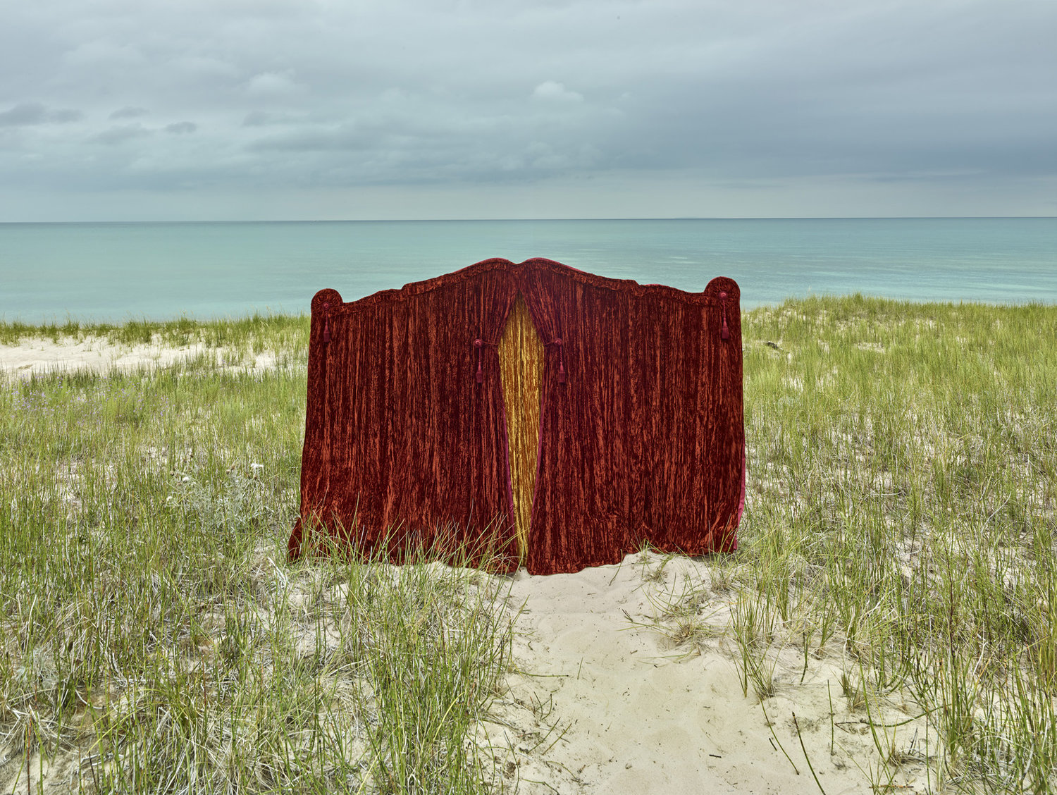 Funeral Homes of the American South by Timothy Hursley — A deep burgundy fabric structure with gold vertical stripe stands isolated on sand dunes amid beach grass, ocean and clouded sky visible beyond.