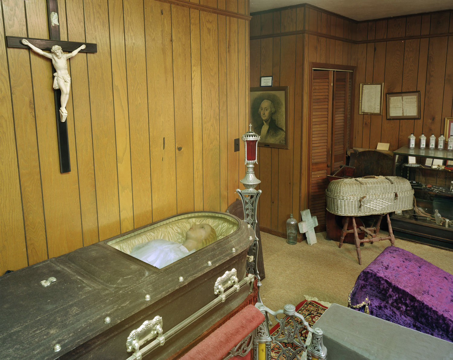 Funeral Homes of the American South by Timothy Hursley — A casket lies open in the foreground of this wood-paneled funeral home chapel, its cream-colored interior stark against dark exterior finish.