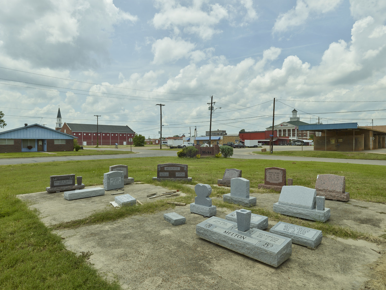 Funeral Homes of the American South by Timothy Hursley — A rural cemetery occupies the foreground of this composition, with granite headstones and markers arranged across concrete pads set into mowed grass.