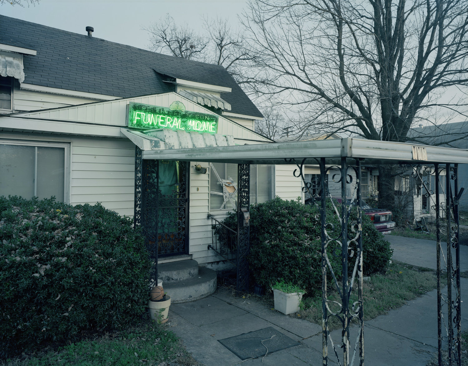 Funeral Homes of the American South by Timothy Hursley — A modest single-story funeral home with horizontal siding and a gabled roof, marked by a bright green neon sign.