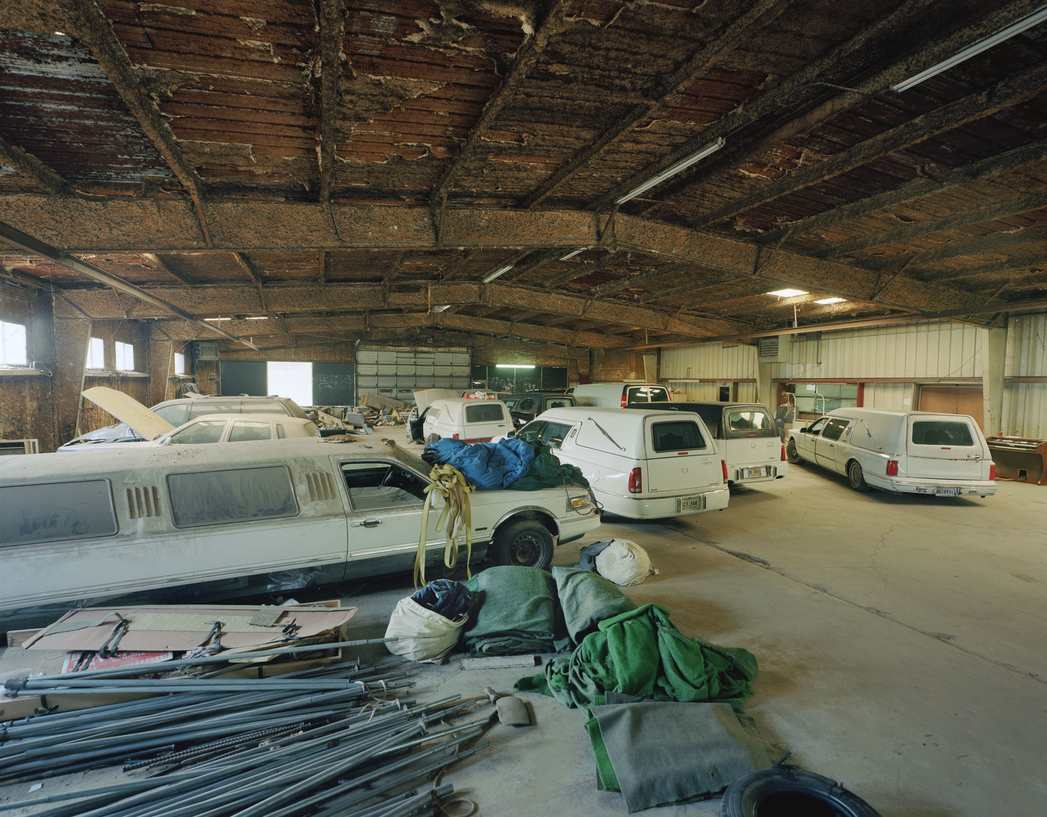 Funeral Homes of the American South by Timothy Hursley — A utilitarian warehouse interior repurposed as storage reveals rows of white and cream-colored hearses and funeral vehicles parked across a concrete floor.
