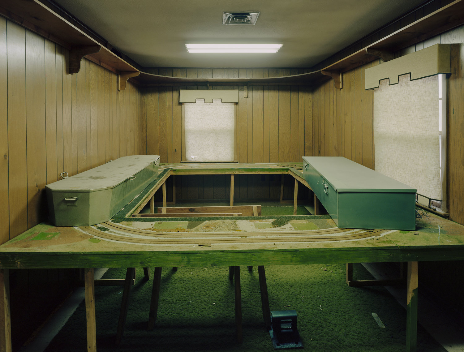 Funeral Homes of the American South by Timothy Hursley — A symmetrical composition of two green caskets positioned on either side of a toy train track that circuits the floor of a wood-paneled viewing room.