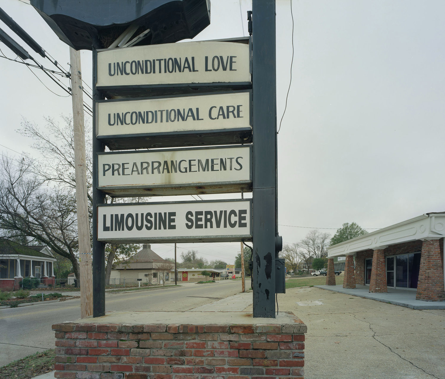 Funeral Homes of the American South by Timothy Hursley — A roadside sign mounted on a steel pole announces funeral services across four horizontal bands: Unconditional Love, Unconditional Care, Prearrangements,...