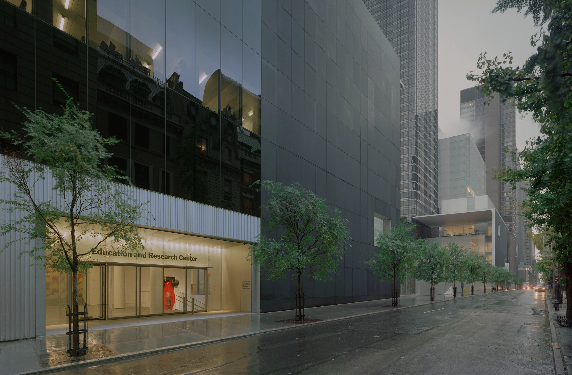 Museum of Modern Art in New York by Yoshio Taniguchi — Street-level view of MoMA's Education and Research Center entrance, characterized by a horizontal band of glass curtain wall with minimal framing set within...