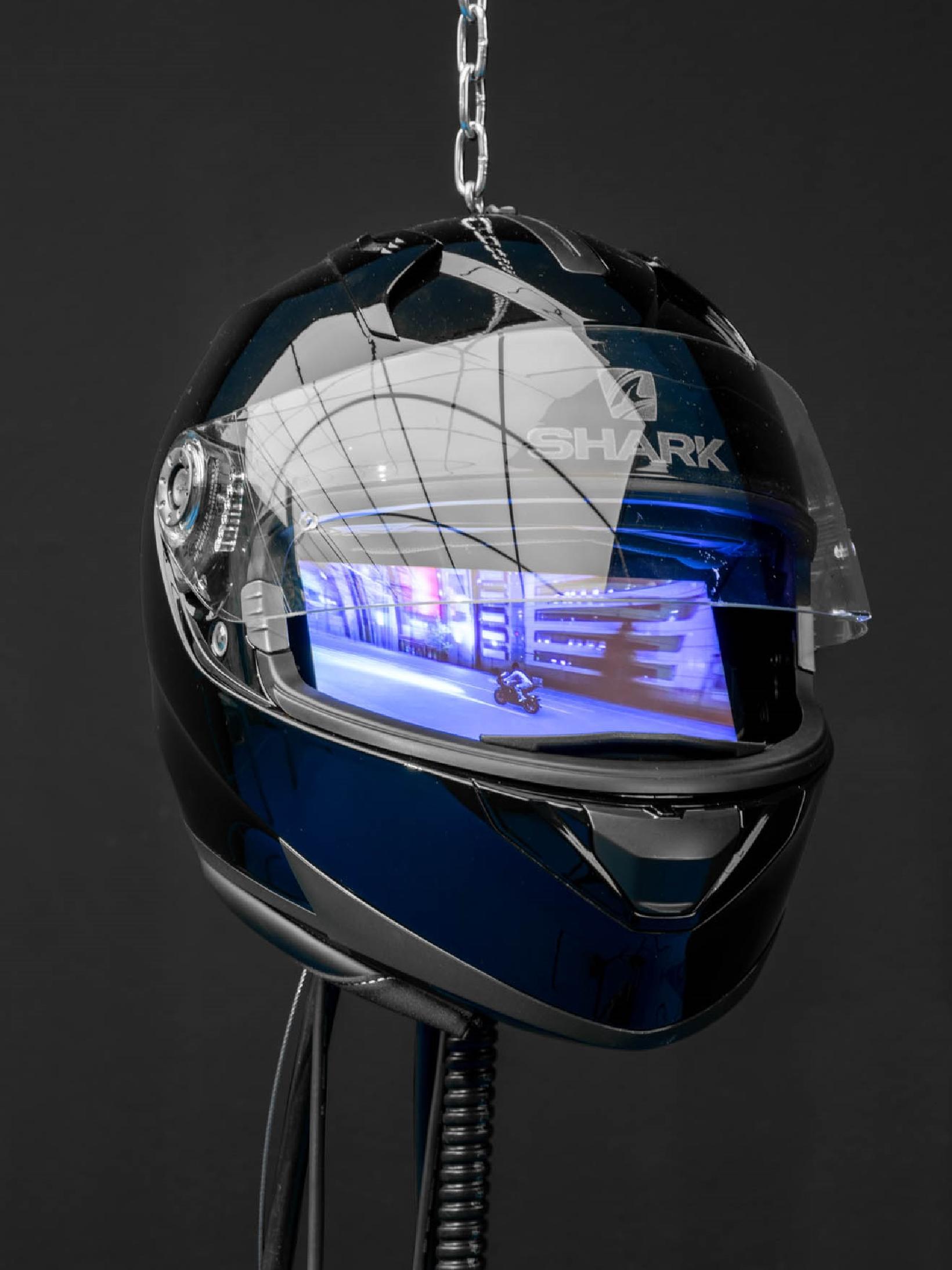 Many Worlds Over by Ayoung Kim at Hamburger Bahnhof — A Shark motorcycle helmet hangs from a silver chain against a dark ground, its visor displaying a rendered interior scene of a modernist corridor bathed in...