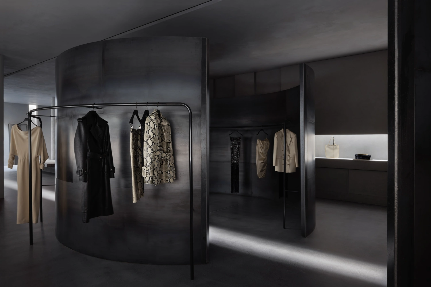 KHAITE Flagship Store by Griffin Frazen — A curved steel partition wall creates a narrowing passage within the flagship store, its dark metallic surface reflecting overhead light.