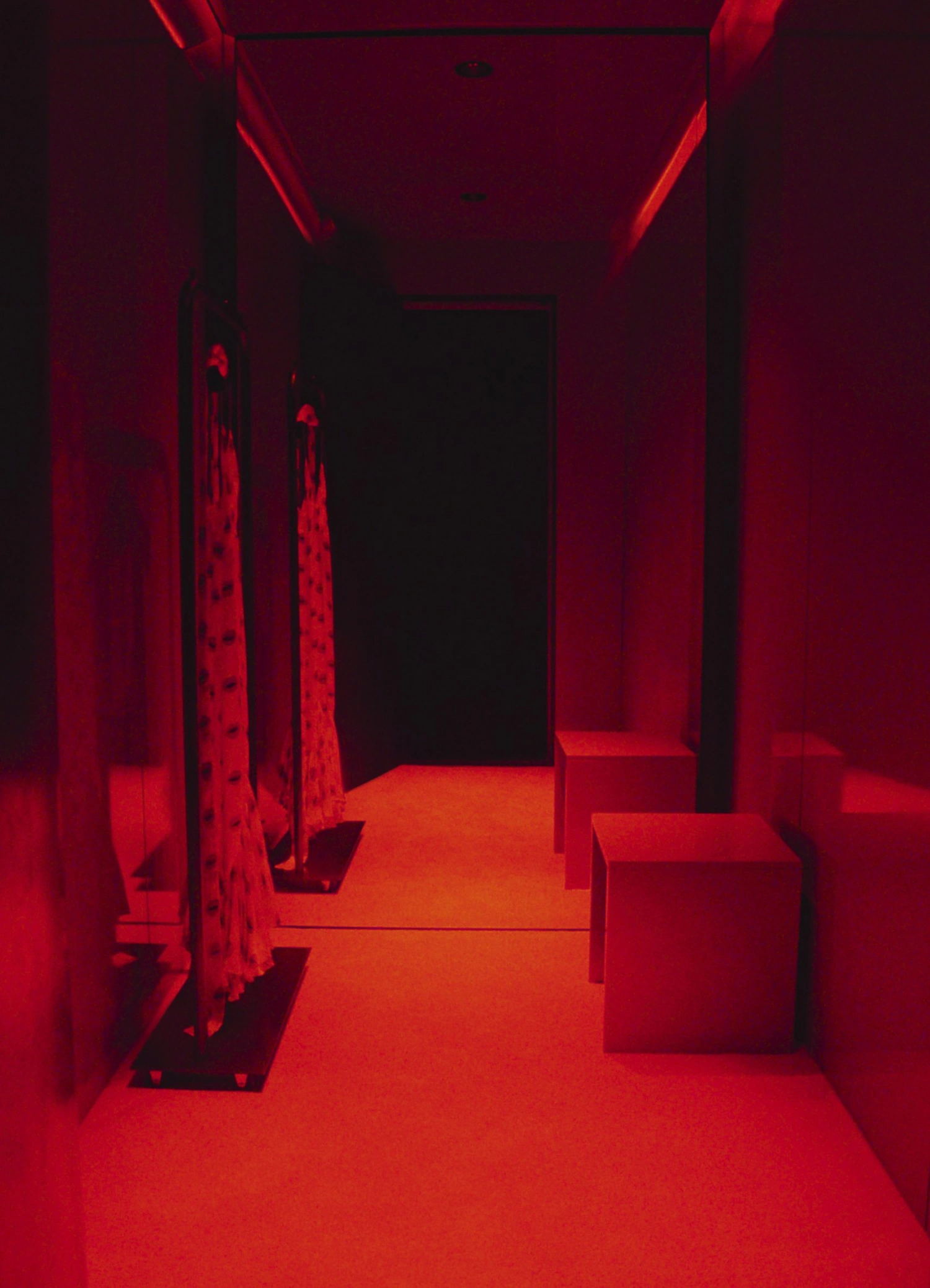 KHAITE Flagship Store by Griffin Frazen — A narrow corridor bathed in red neon light, its polished concrete floor transitioning from deep crimson to warm orange as it recedes toward a darkened doorway.