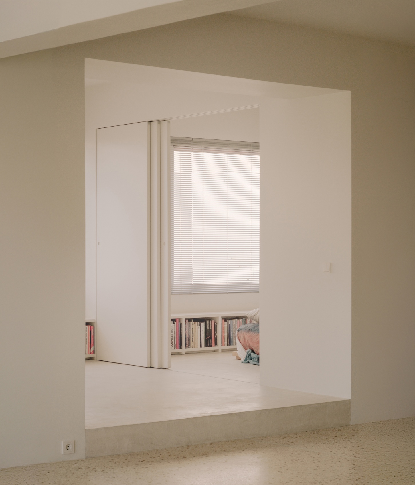 'Astral Weeks' by Oblique — A recessed alcove carved into the perimeter wall reveals a secondary living zone with built-in shelving beneath a horizontal window fitted with venetian blinds.