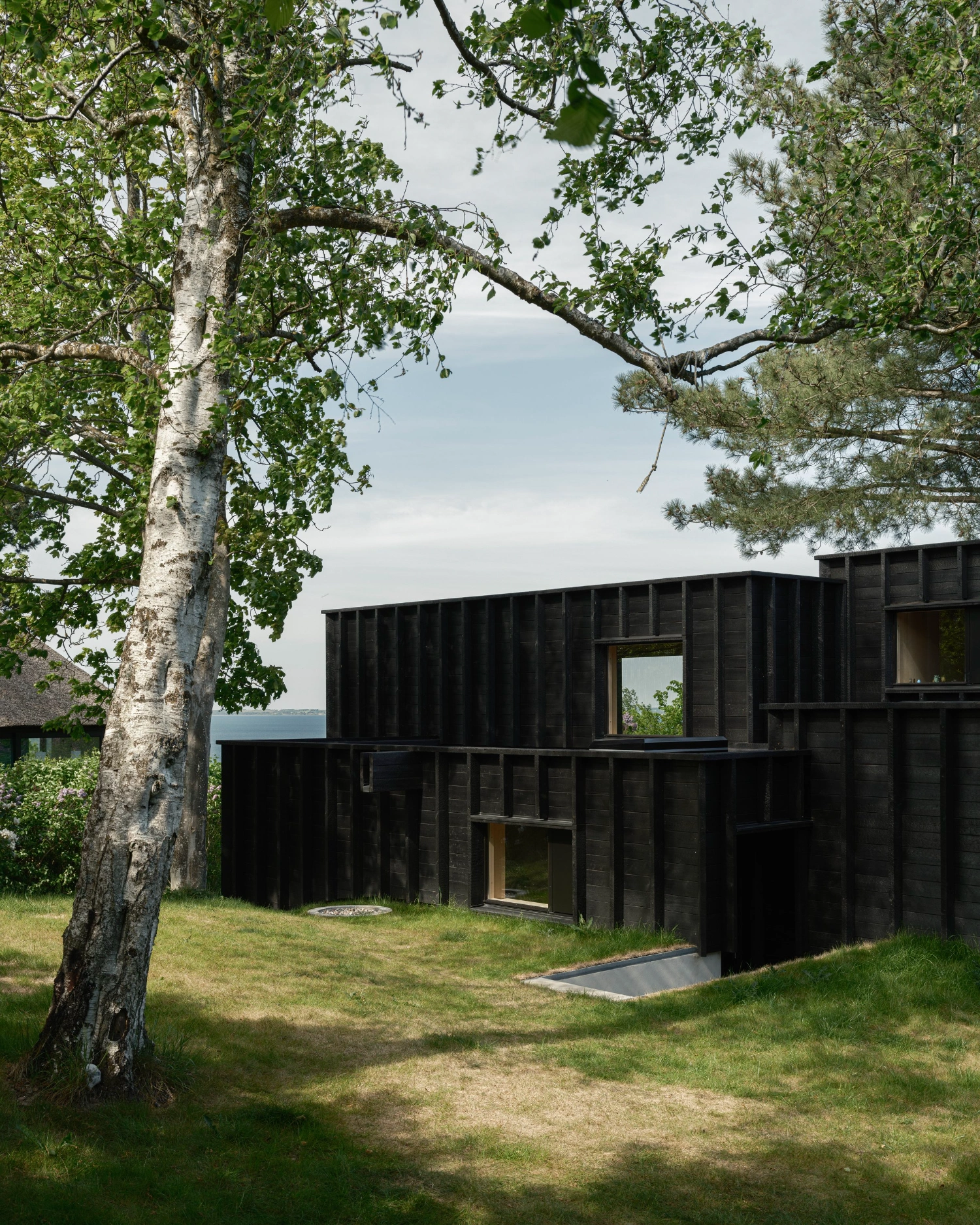 House with a Hidden Atrium by Förstberg Ling — A blackened timber-clad extension emerges from a coastal landscape, its facade composed of vertical pine ribs casting deep shadow lines across the dark surface.