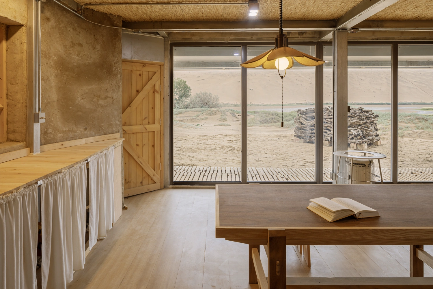 Home in the Badain Jaran Desert by School of Architecture and Urban Planning NJU — Interior view of rammed-earth dwelling featuring a timber table with integrated storage and woven rush seating arranged in pairs.