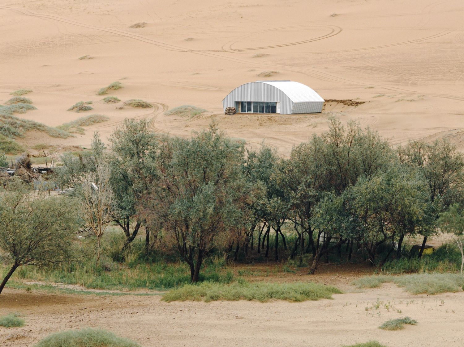 Home in the Badain Jaran Desert by School of Architecture and Urban Planning NJU — A corrugated metal and white-paneled warehouse structure sits isolated on the sandy plain of the Badain Jaran Desert, its arched roof catching afternoon light.