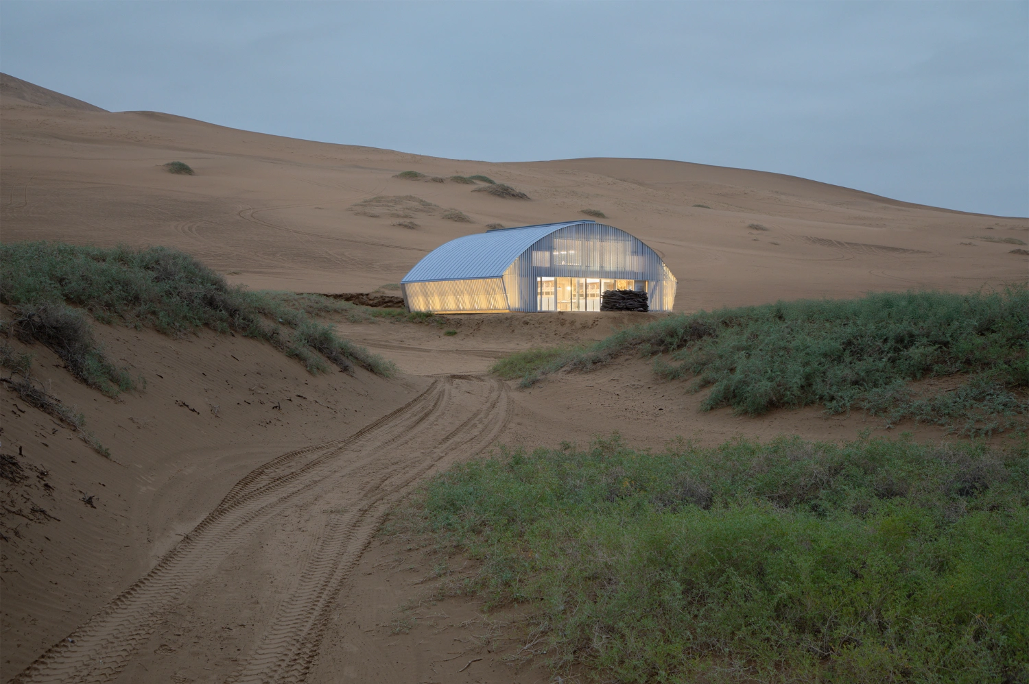 Home in the Badain Jaran Desert by School of Architecture and Urban Planning NJU — A corrugated-metal structure with a barrel-vault roof sits embedded in sandy terrain, its white panels glowing from interior illumination against the dusk sky.