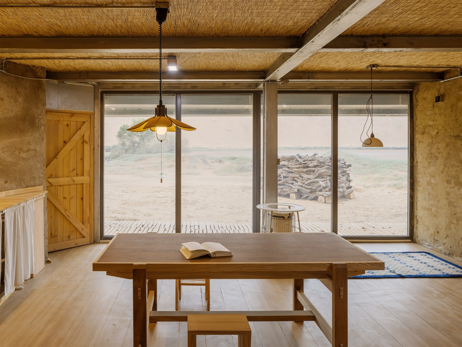 Home in the Badain Jaran Desert by School of Architecture and Urban Planning NJU — A timber table dominates the central volume, its broad surface anchoring the open-plan interior beneath a skylight roof with exposed metal cross-bracing and...
