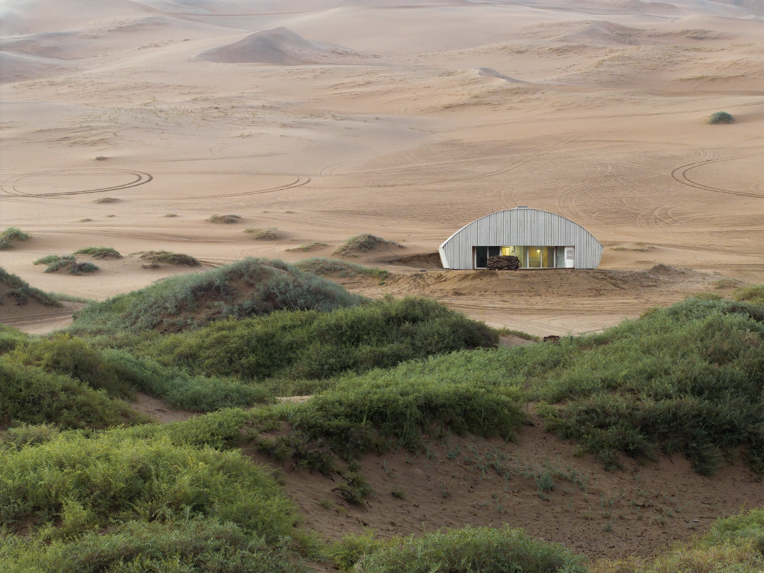 Home in the Badain Jaran Desert by School of Architecture and Urban Planning NJU — A corrugated metal structure with a distinctive arched roof sits embedded in the desert landscape, its white-painted surface a deliberate counterpoint to...