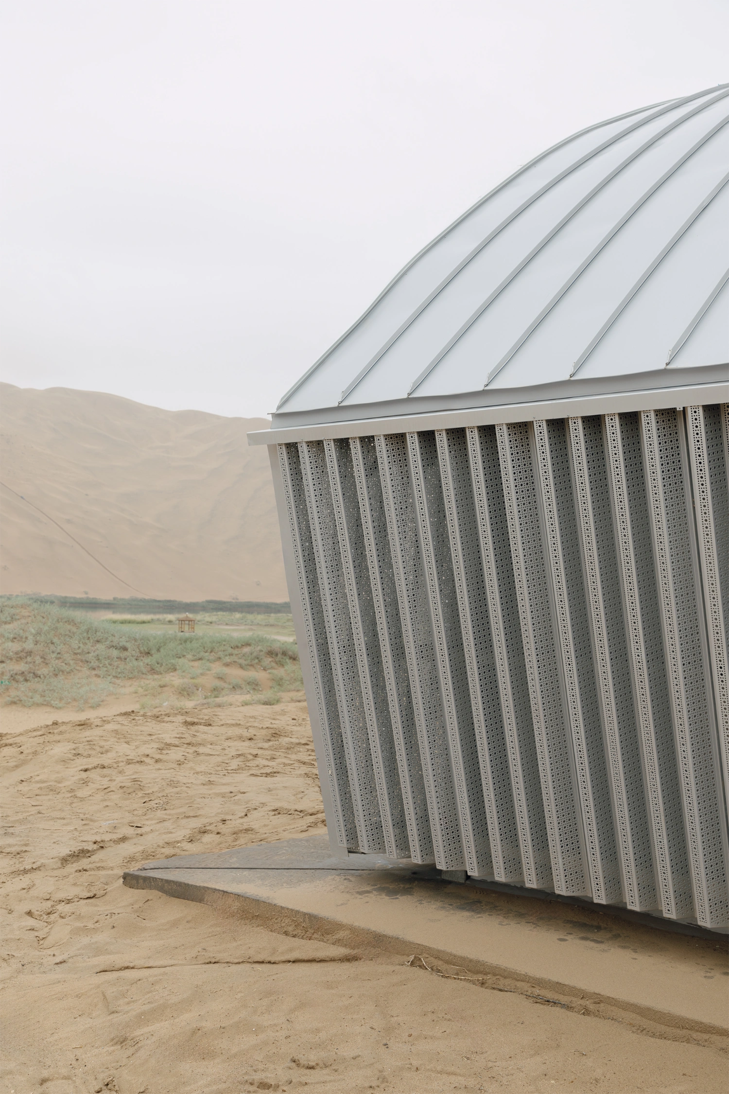 Home in the Badain Jaran Desert by School of Architecture and Urban Planning NJU — A corrugated metal perforated screen wall dominates the foreground, its vertical linear ridges catching diffuse light against a pale sky.