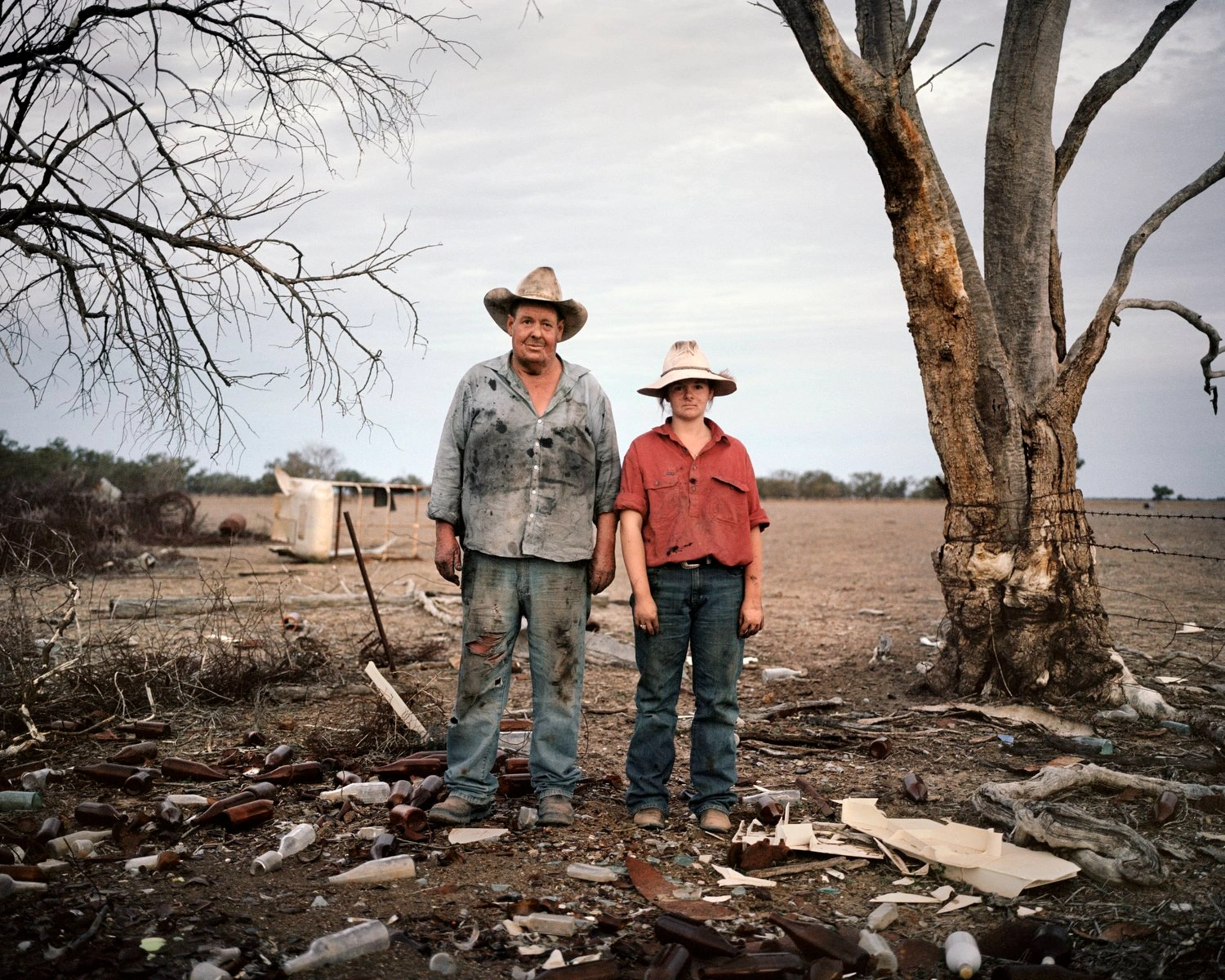 Big Sky by Adam Ferguson — Two figures stand beneath a gnarled, bare-branched tree in arid scrubland, their worn work clothes and wide-brimmed hats marking them as rural laborers.