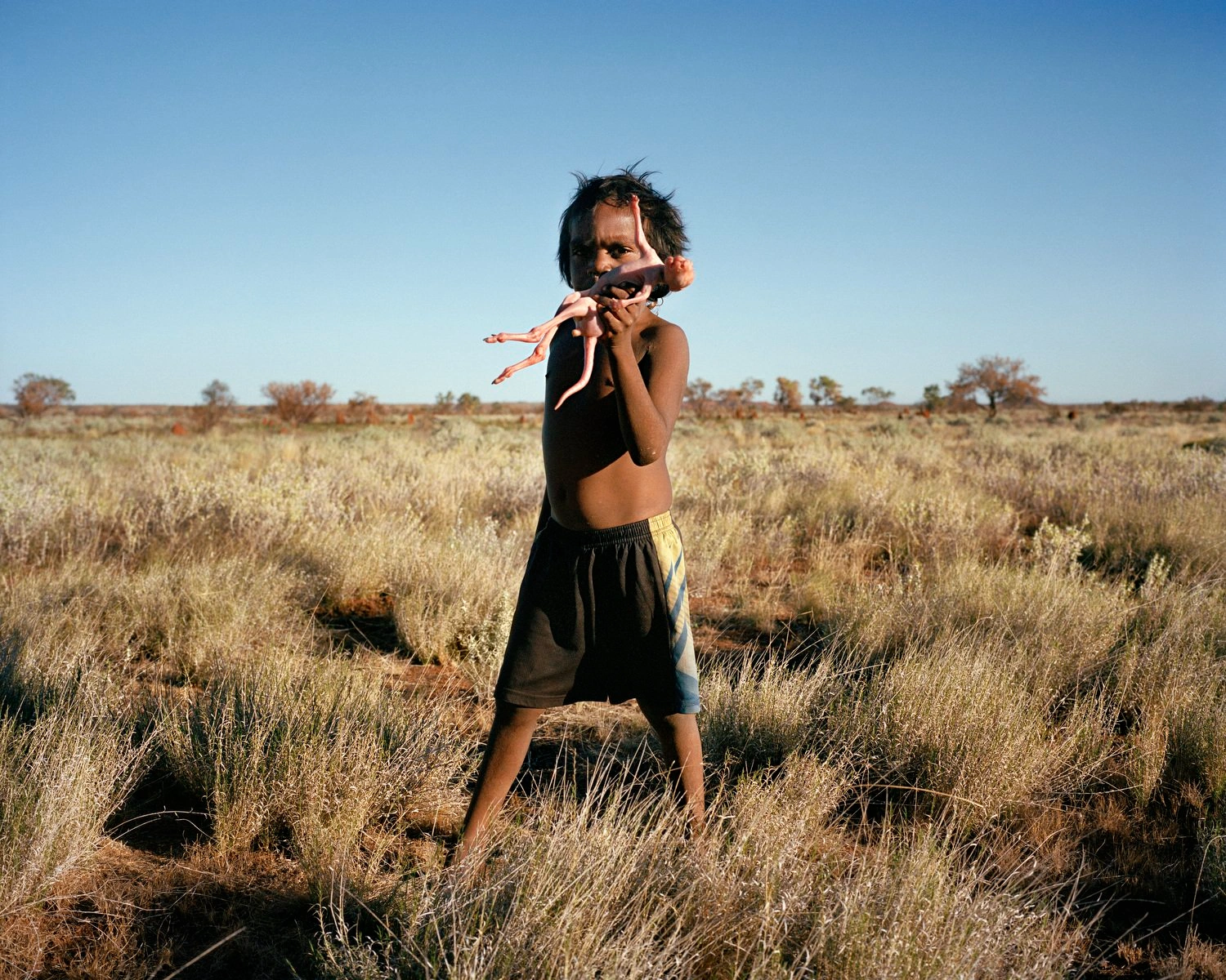Big Sky by Adam Ferguson — A young figure stands centrally in expansive Australian scrubland, arms raised in an open gesture against a clear sky.