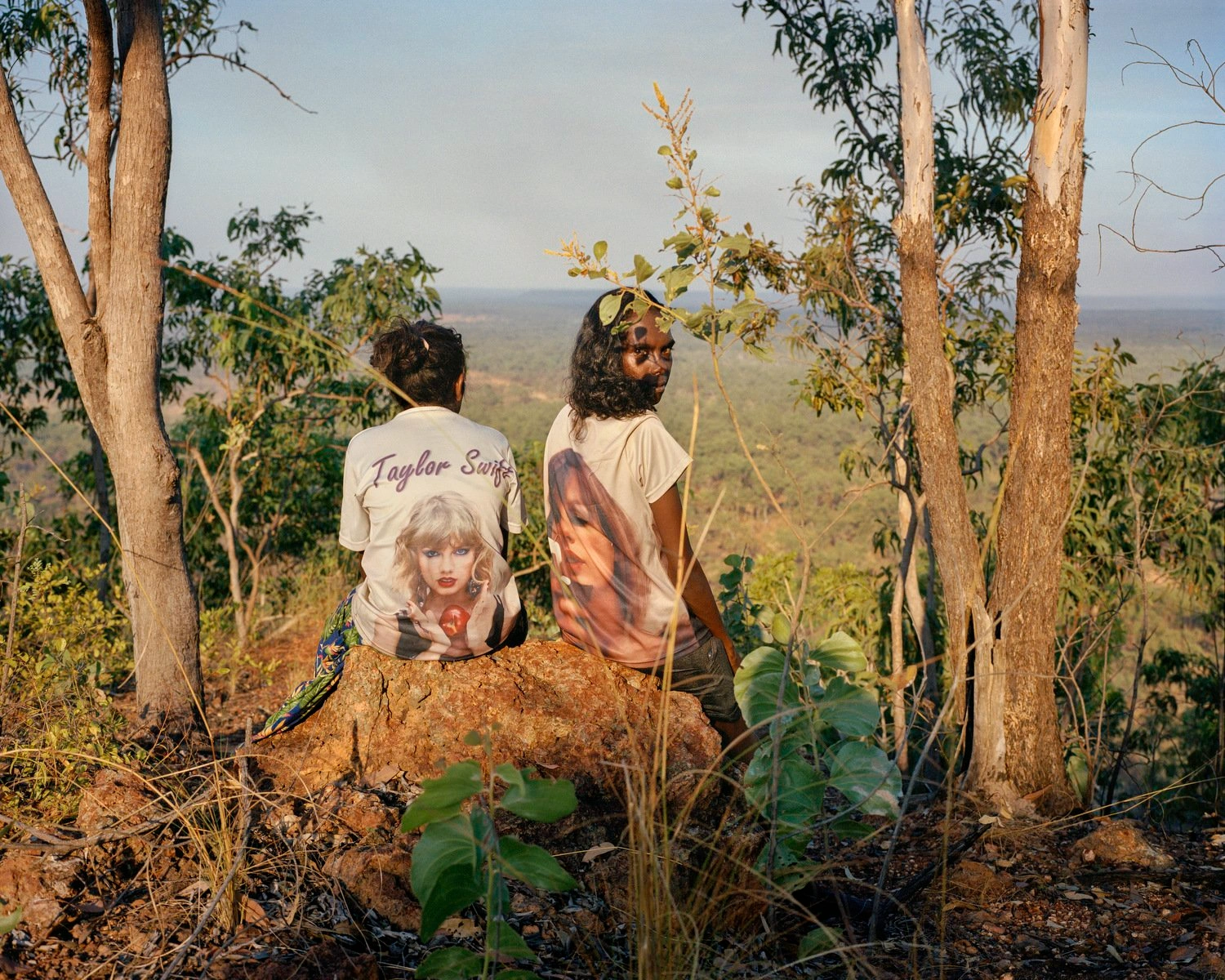 Big Sky by Adam Ferguson — Two figures sit on exposed reddish earth among eucalyptus trees, their backs to camera, facing an expansive landscape of undulating hills under clear sky.