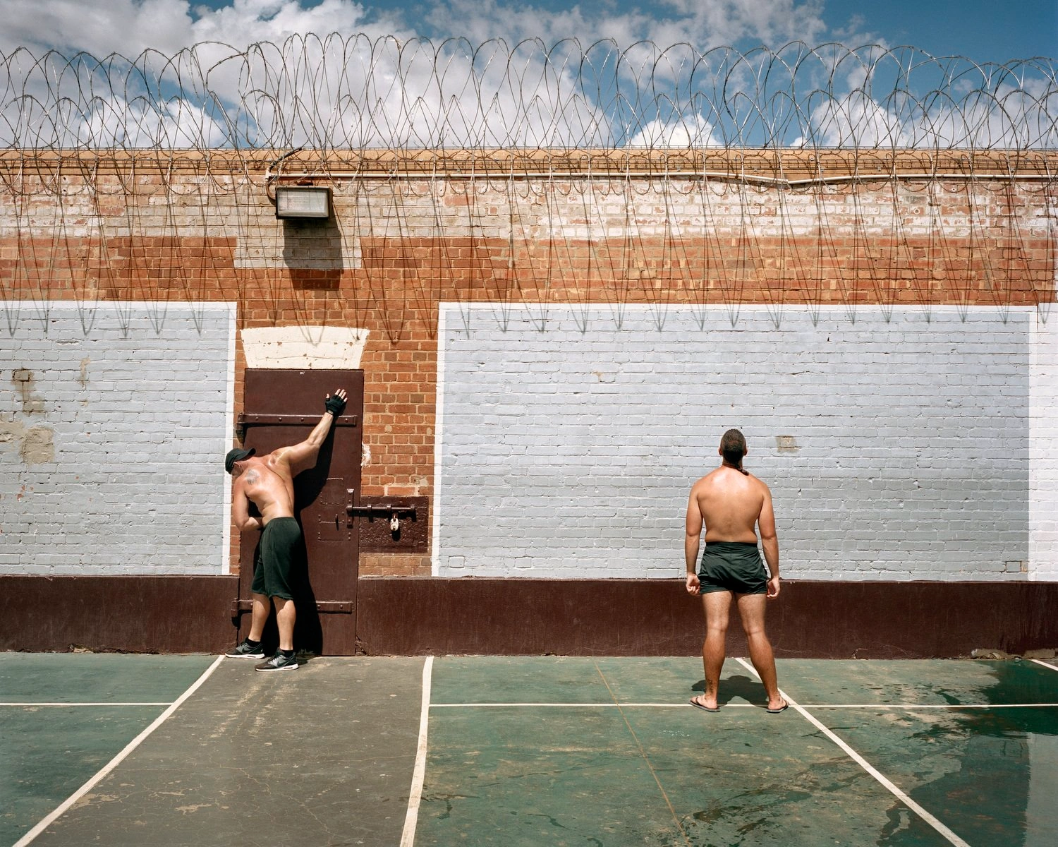 Big Sky by Adam Ferguson — Two shirtless men occupy a deteriorating outdoor court bounded by a brick structure topped with rusted barbed wire.