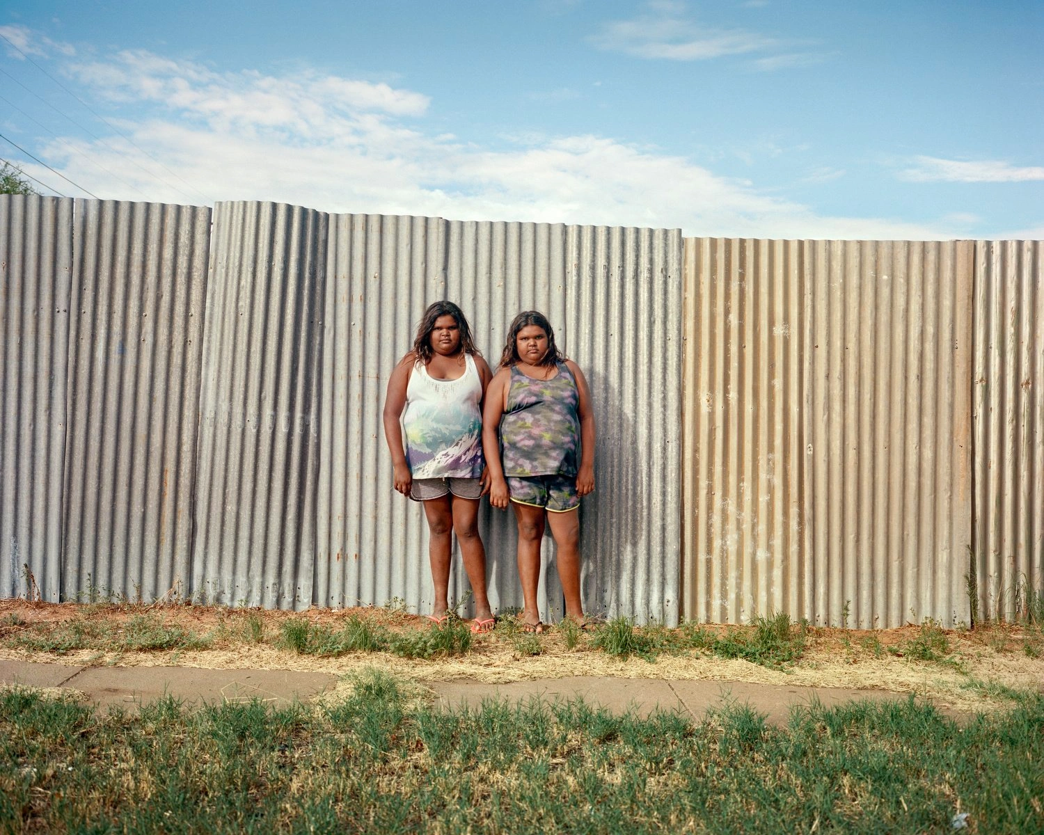 Big Sky by Adam Ferguson — Two figures stand centered against a corrugated metal fence, its weathered galvanized surface showing distinct color variation from grey-silver on the left...