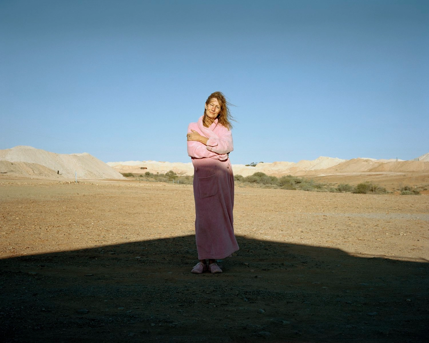 Big Sky by Adam Ferguson — A woman stands centrally in a vast Australian desert landscape, wearing a pink linen shirt and burgundy skirt that contrasts sharply against the exposed earth.