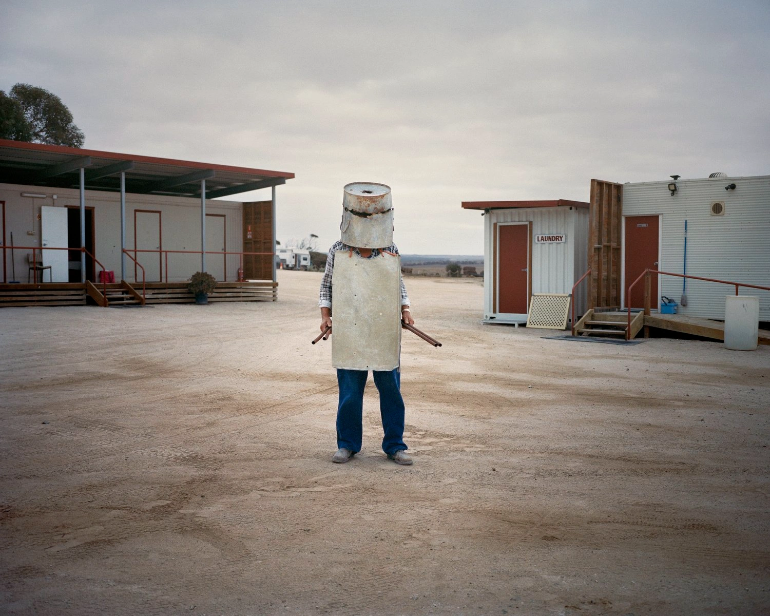 Big Sky by Adam Ferguson — A figure stands centrally in an expansive desert settlement, their head obscured by a weathered cylindrical concrete structure—likely salvaged industrial or...