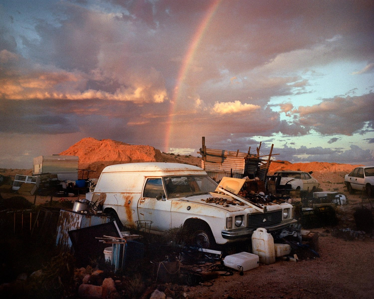 Big Sky by Adam Ferguson — A rusted and dismantled white sedan sits exposed in an arid Australian junkyard, its hood propped open to reveal stripped engine components.