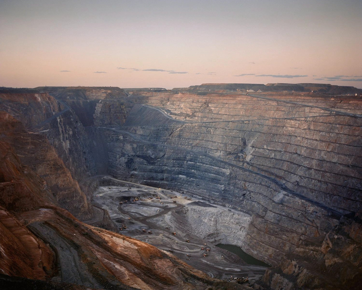 Big Sky by Adam Ferguson — An open-pit mining operation photographed at dusk reveals concentric terraced walls of exposed stone and earth in shades of rust, ochre, and grey.
