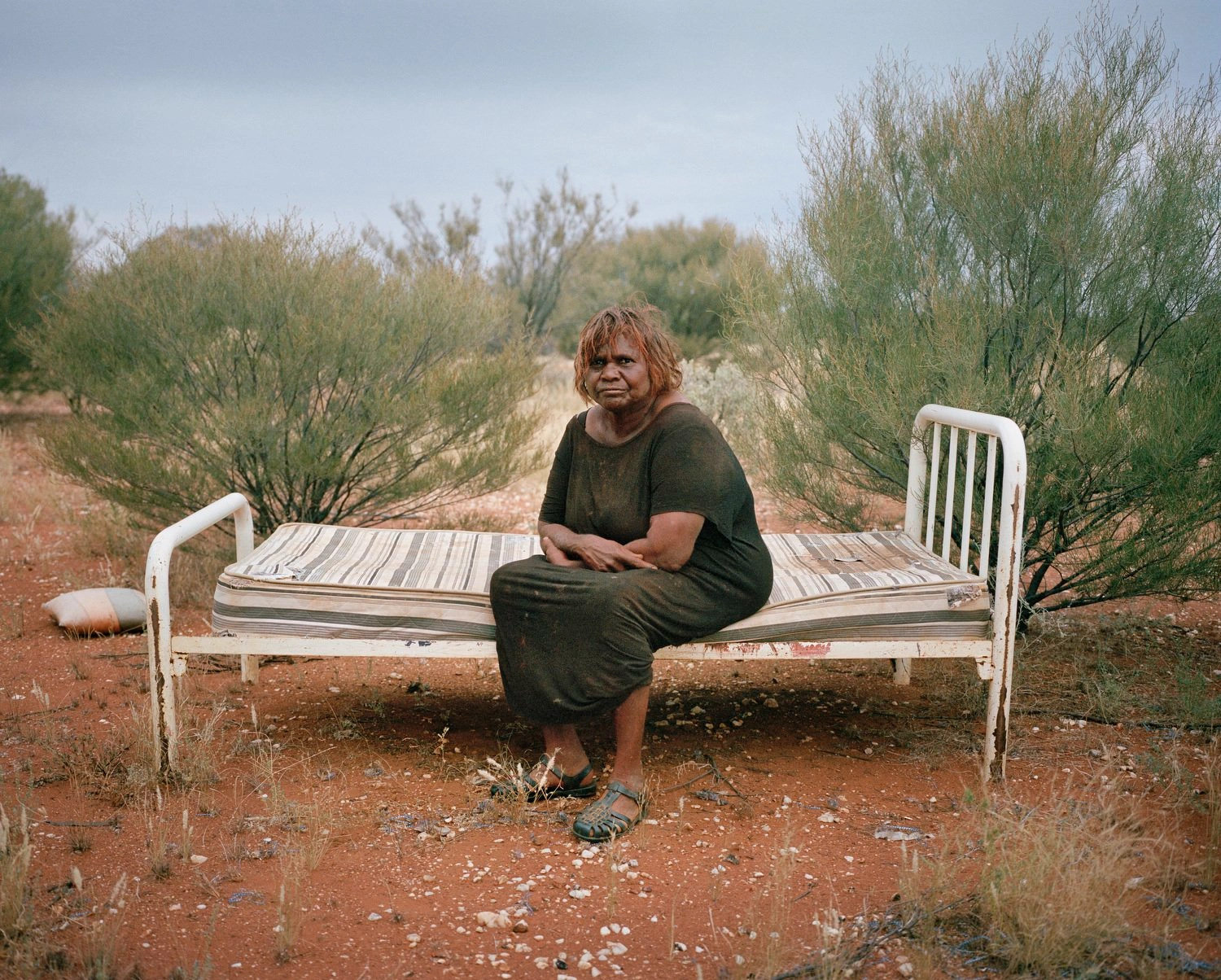 Big Sky by Adam Ferguson — A figure in olive-green cotton sits centrally on a white metal-framed bed positioned in sparse Australian scrubland.