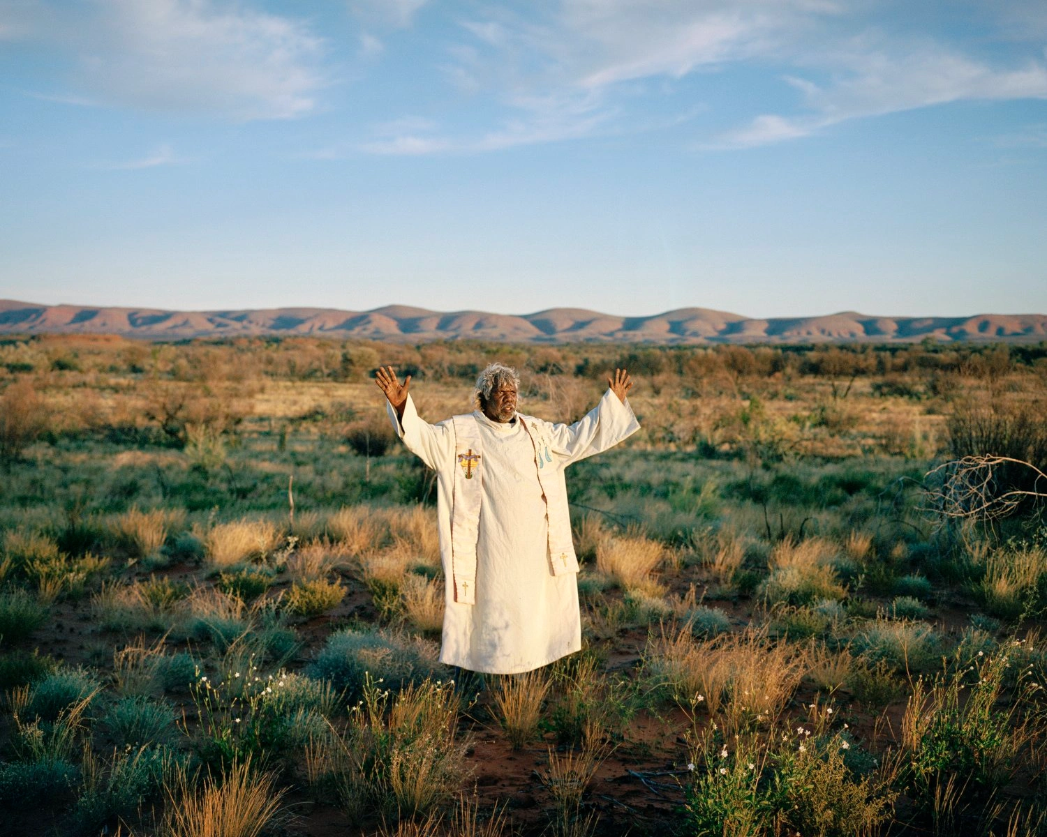Big Sky by Adam Ferguson — A figure in cream linen stands centrally in arid scrubland, arms raised in an open gesture against a vast desert landscape.
