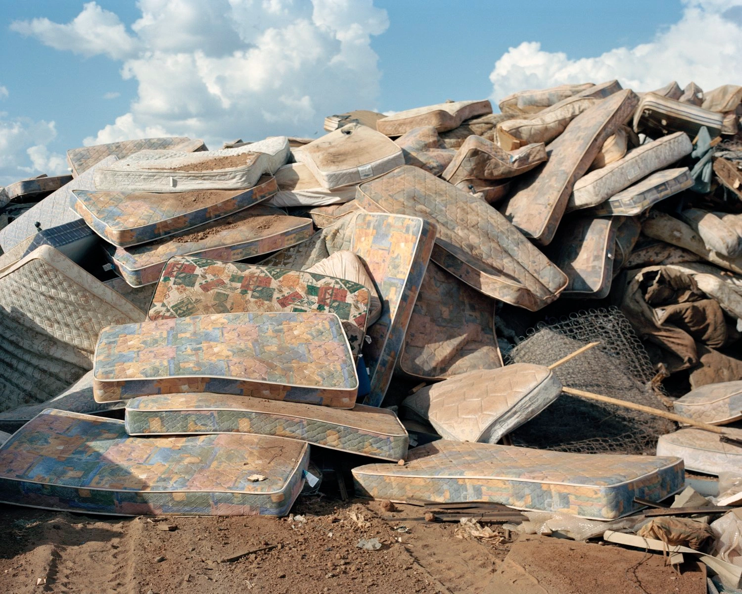 Big Sky by Adam Ferguson — A sprawling pile of discarded mattresses accumulates in the Australian outback, their worn fabrics—patterned in faded florals, stripes, and geometric...