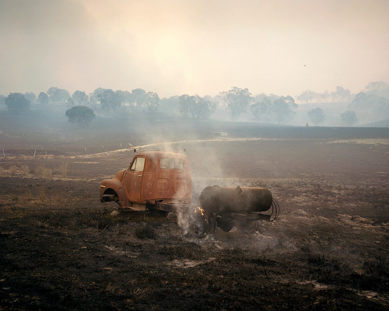 Big Sky by Adam Ferguson — A rusted orange truck and attached water tank sit abandoned in scorched earth, actively releasing steam into hazy air.