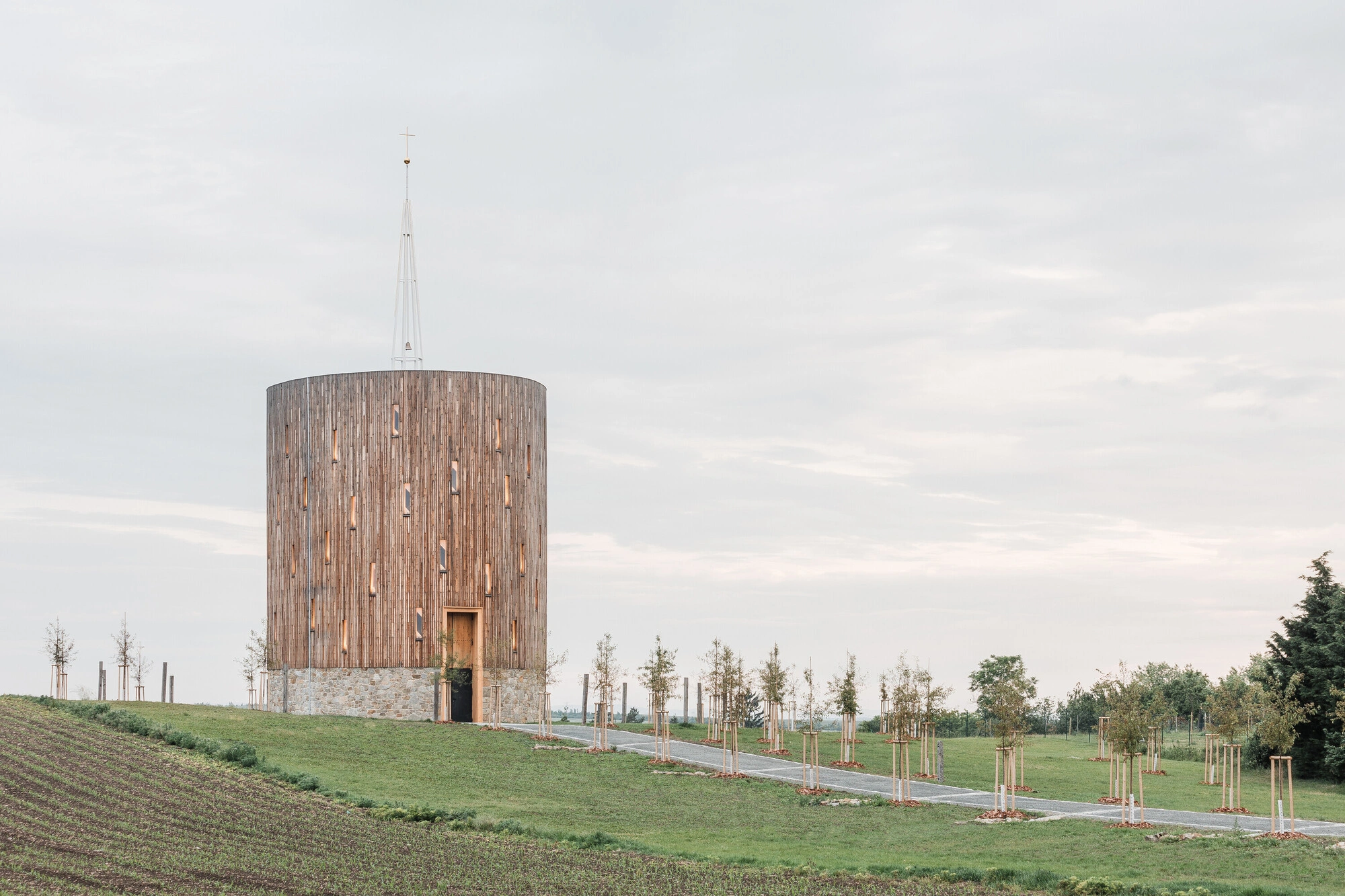 Our Lady of Sorrows Chapel by RCNKSK — The cylindrical timber-clad chapel dominates a gently sloped pastoral landscape, its vertical wood boards creating pronounced linear striations across the...