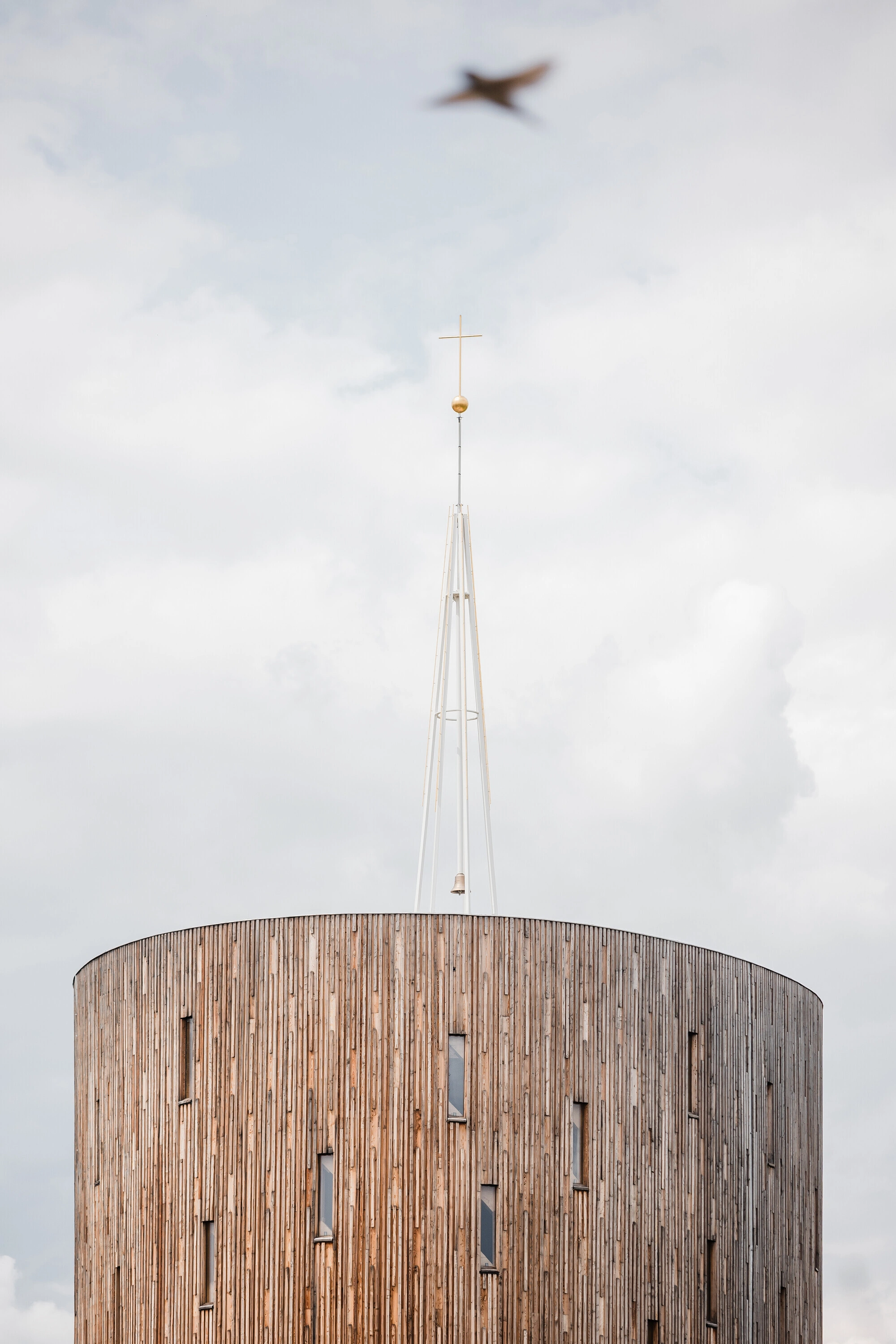 Our Lady of Sorrows Chapel by RCNKSK — The chapel's cylindrical timber-clad volume rises against overcast sky, its vertical board cladding emphasizing height toward a slender white spire topped...